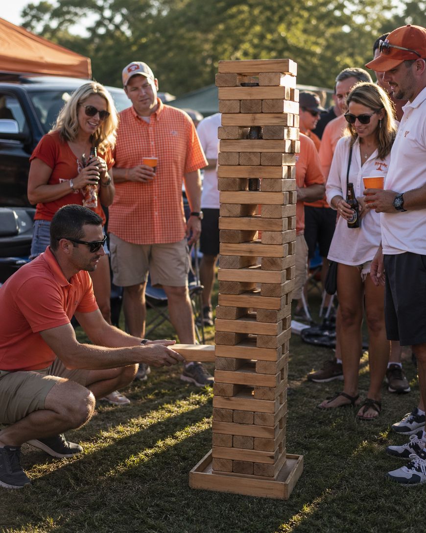 A group of people outdoors watch as a man kneels to carefully pull a block from a tall wooden Jenga tower. Most are wearing casual summer clothes and sunglasses. The sun is shining and there are trees in the background.