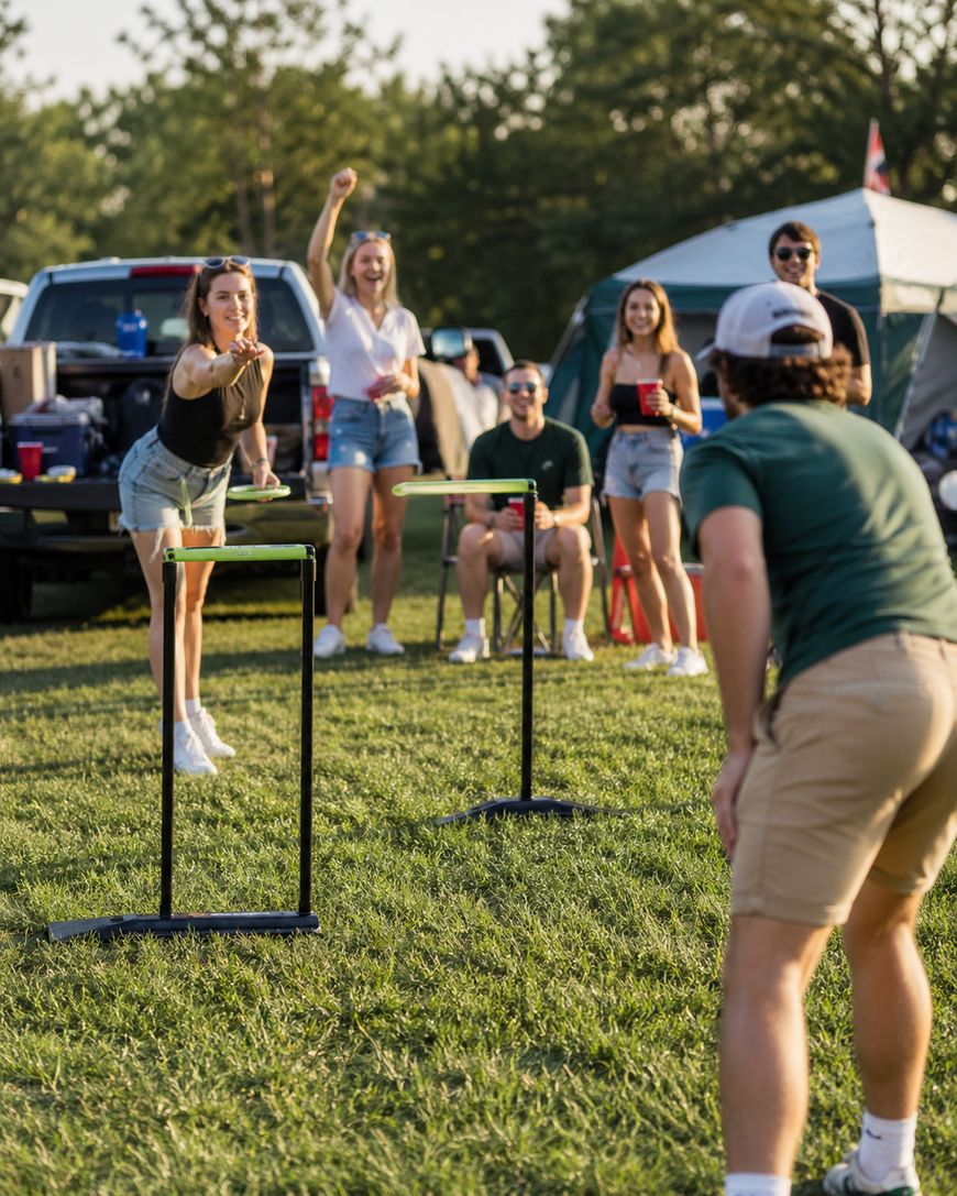 A group of young adults play an outdoor game on grass. A woman throws a frisbee towards a man; others watch and smile. Theres a pickup truck, tent, and trees in the background, suggesting a casual, sunny gathering.