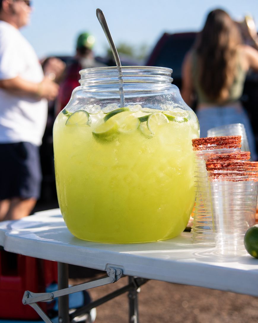 A large glass jar filled with lime slices and a green beverage sits on a white table next to a stack of clear plastic cups rimmed with chili powder; people and cars are blurred in the background.