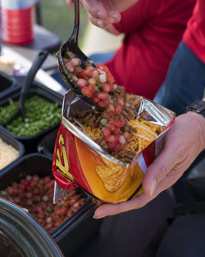 A person scoops salsa onto a bag of corn chips filled with ground beef and shredded cheese, preparing walking tacos at an outdoor food station with various toppings in containers.