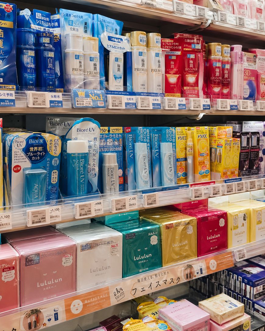 Shelves in a Japanese store displaying various skincare and beauty products in colorful packaging, including Biore UV sunscreen and Lululun face masks. Labels and signs are mostly in Japanese.