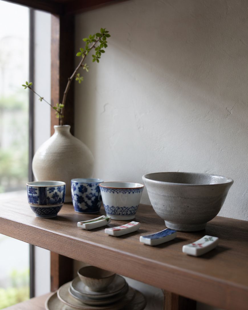 Ceramic cups, bowls, and a vase with green sprigs are arranged neatly on wooden shelves by a window, with sunlight streaming in. Four decorative porcelain chopstick rests are displayed on the lower shelf.
