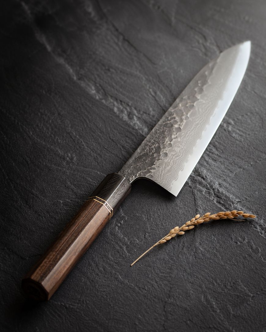 A Japanese chef knife with a dark wooden handle and a textured, patterned blade rests on a dark stone surface next to a single stalk of wheat.