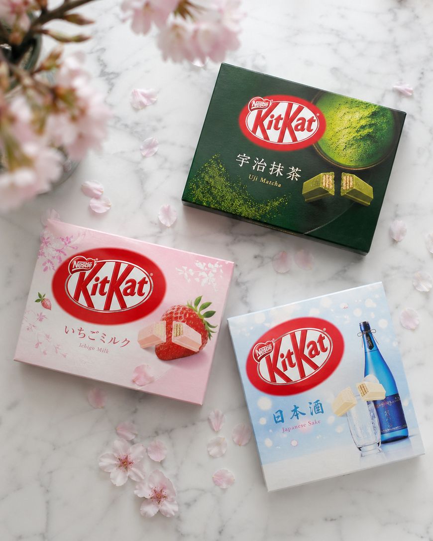 Three boxes of Japanese Kit Kat flavors—Uji Matcha (green tea), Sakura Milk (pink), and Japanese Sake (white)—are arranged on a marble surface with scattered cherry blossom petals and flowers.