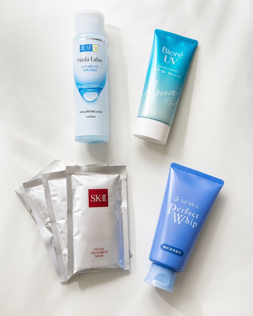 Four skincare products on a white surface: a Hada Labo lotion bottle, a Biore UV Aqua Rich sunscreen tube, a blue Senka Perfect Whip tube, and three silver SK-II facial treatment mask packets.