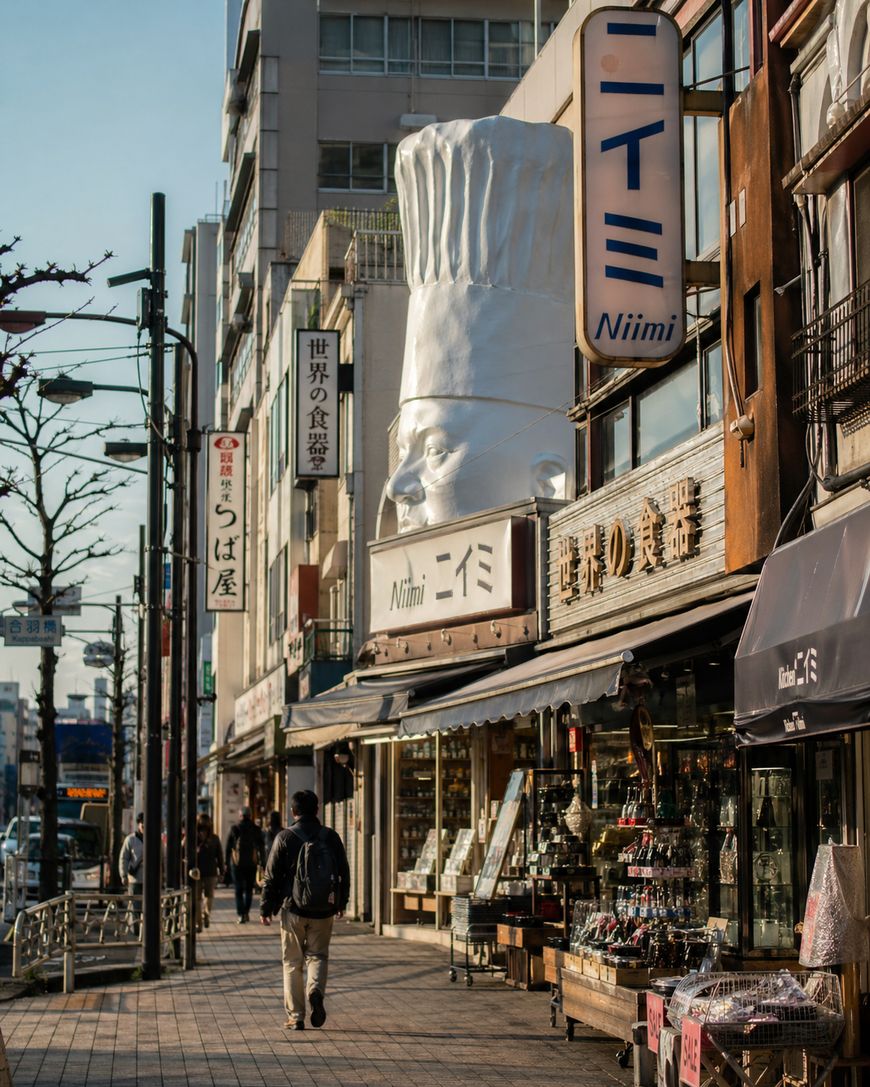 A city street in Japan features shops with Japanese signage and a large sculpture of a chef’s head wearing a tall hat on top of a building. People walk along the sidewalk on a sunny day.