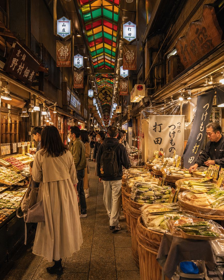 People browse stalls filled with food and goods inside a lively, well-lit Japanese market with colorful stained glass ceiling panels and hanging lantern signs. The atmosphere is bustling and inviting.