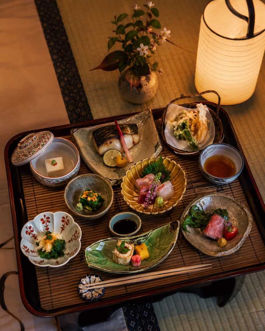 A Japanese kaiseki meal is beautifully arranged on a tray with various small dishes of sashimi, grilled fish, tofu, pickles, vegetables, dipping sauces, and a decorative lamp in the background.
