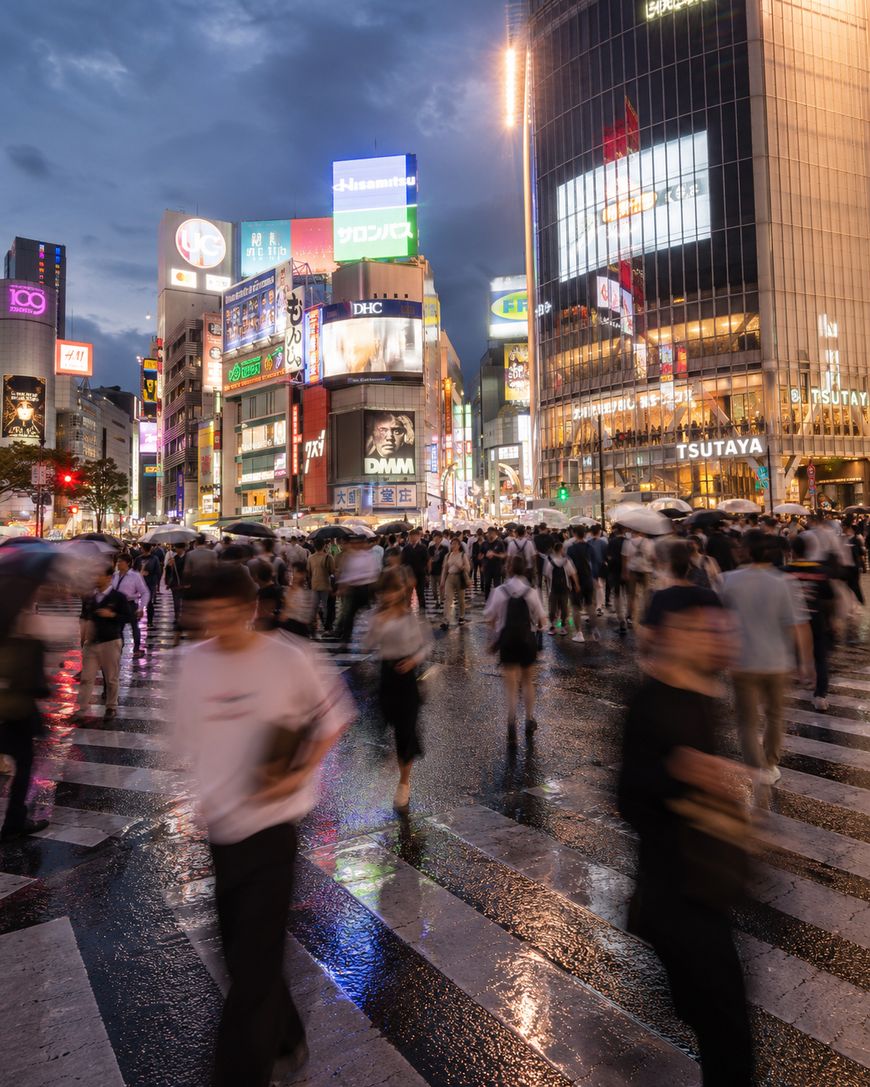 Crowds of people cross a busy intersection in a city at dusk, with bright neon signs and large buildings illuminated in the background. The wet street reflects the lights, and some people appear blurred in motion.
