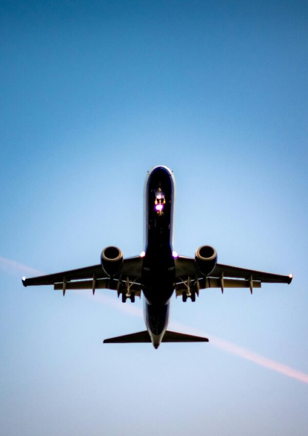A commercial airplane is flying overhead against a clear blue sky, viewed from directly below. The jets landing gear is up, and lights are visible on the front of the aircraft.
