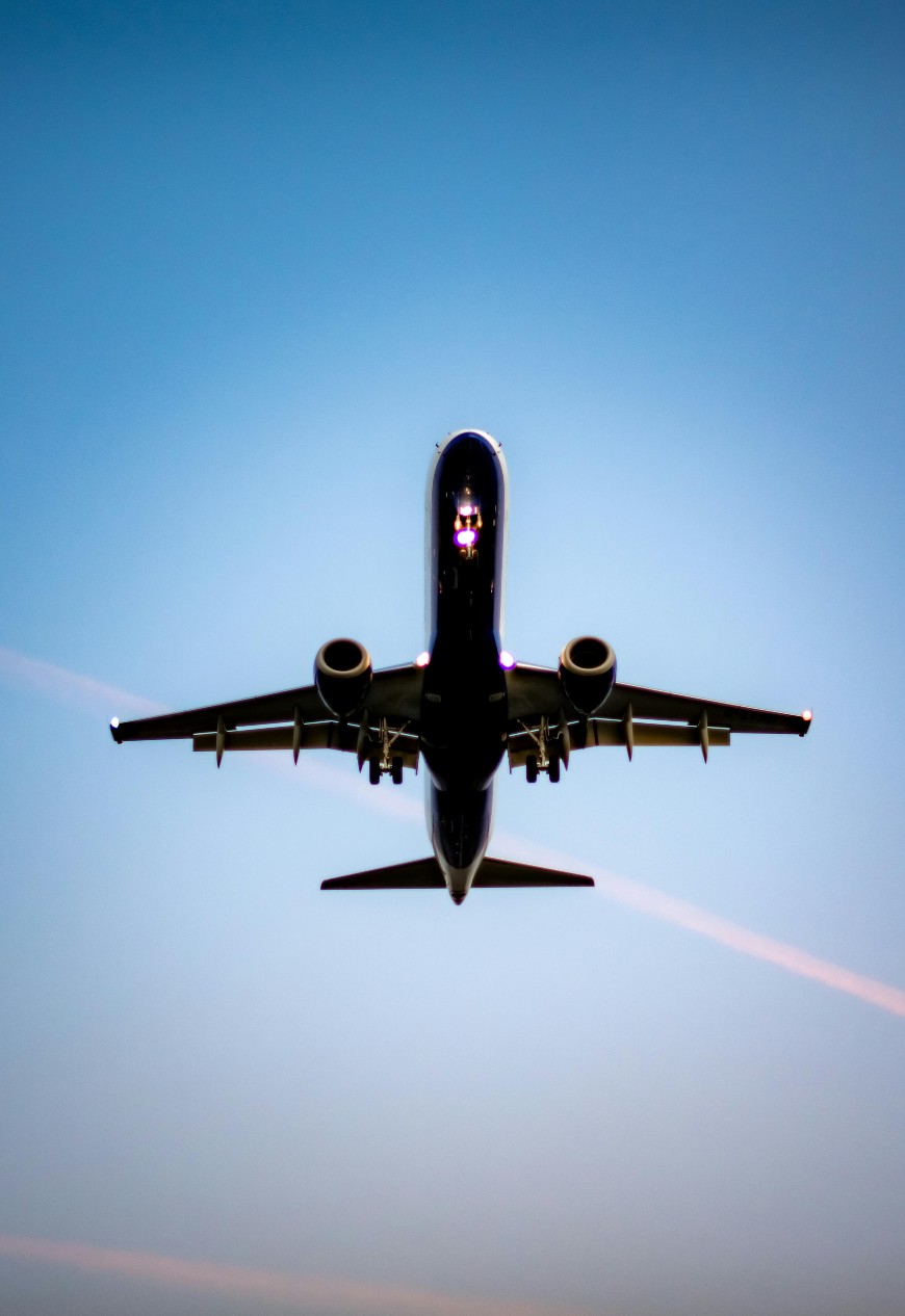 A commercial airplane is seen from below, flying against a clear blue sky with faint contrails in the background. The aircrafts lights and engines are visible, and it appears to be in mid-flight.