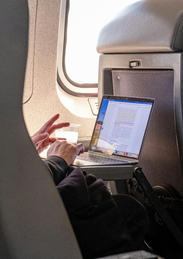A person works on a laptop with a highlighted document open, seated by an airplane window. A small cup is held in one hand, while the laptop rests on the tray table in front of them.