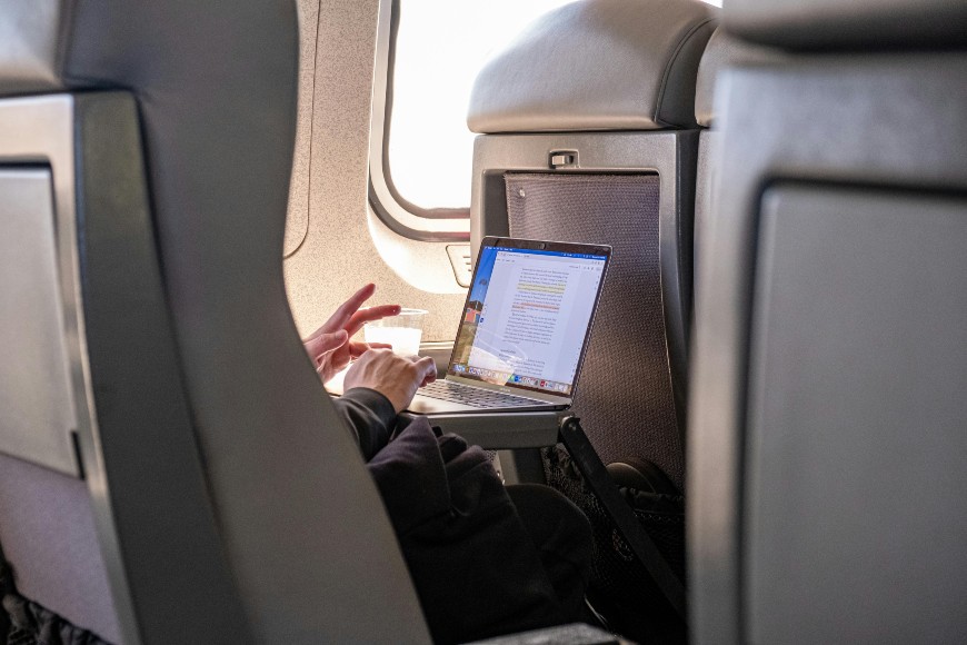 A person works on a laptop at a tray table while flying on an airplane, holding a cup in one hand. The cabin seats and a window with daylight are visible.