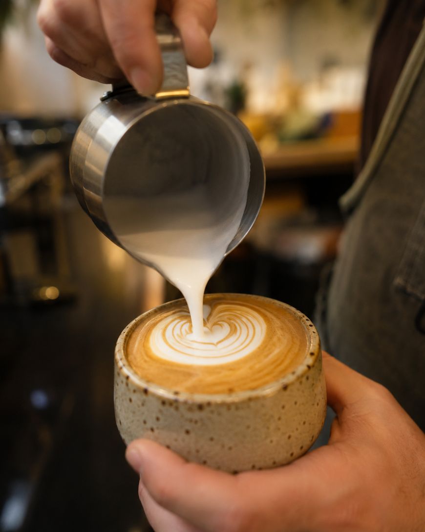 A person pours steamed milk from a metal pitcher into a cup of coffee, creating a latte art pattern on the surface. The hands and cup are in focus, with a blurred background of a cafe.