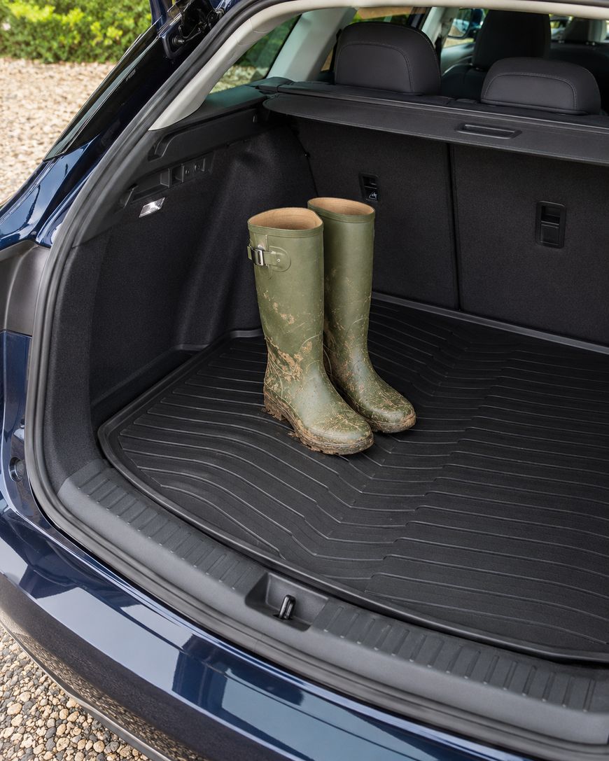 A pair of muddy green rubber boots stands in the cargo area of a car with the trunk open, showing a clean, black, all-weather cargo mat.
