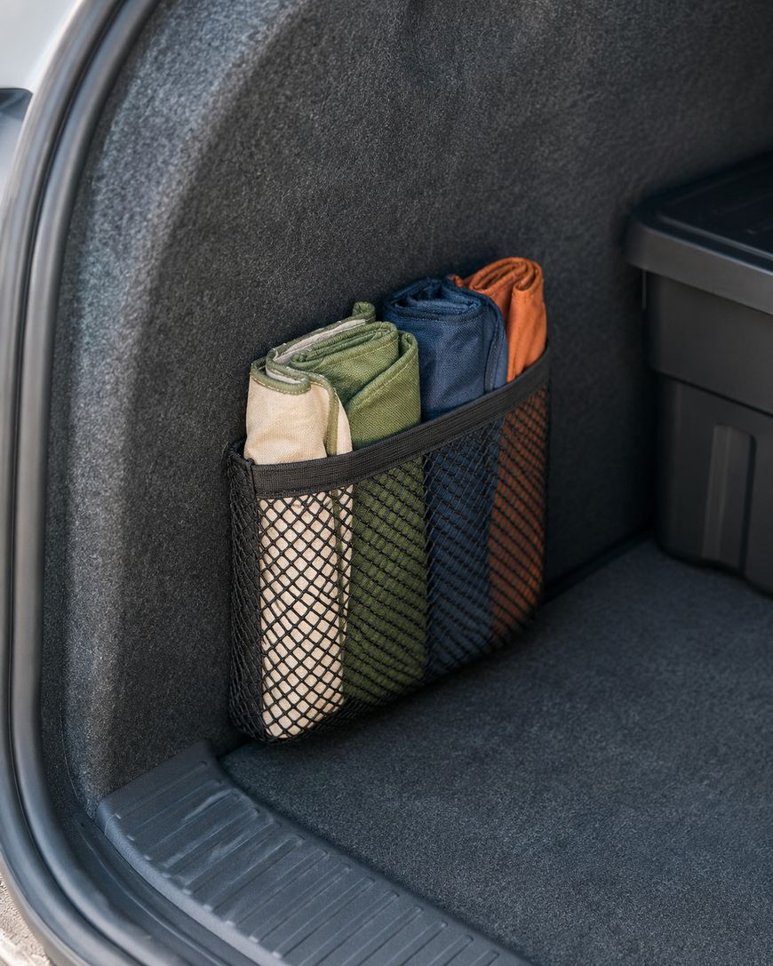 A car trunk with a black mesh pocket holding three neatly folded fabric items in green, blue, and orange, next to a black plastic storage container.