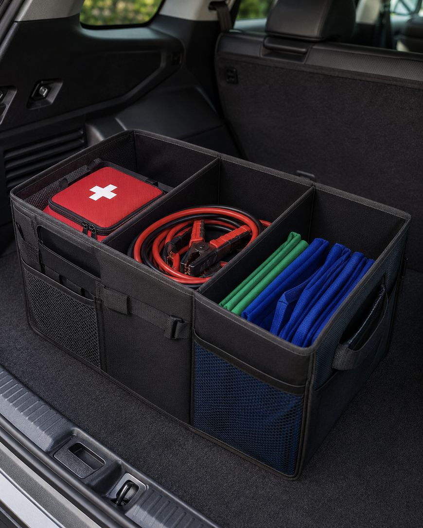 A black car trunk organizer holds a red first aid kit, jumper cables, green and blue folded cloths, and other supplies, placed in the back of a vehicle.