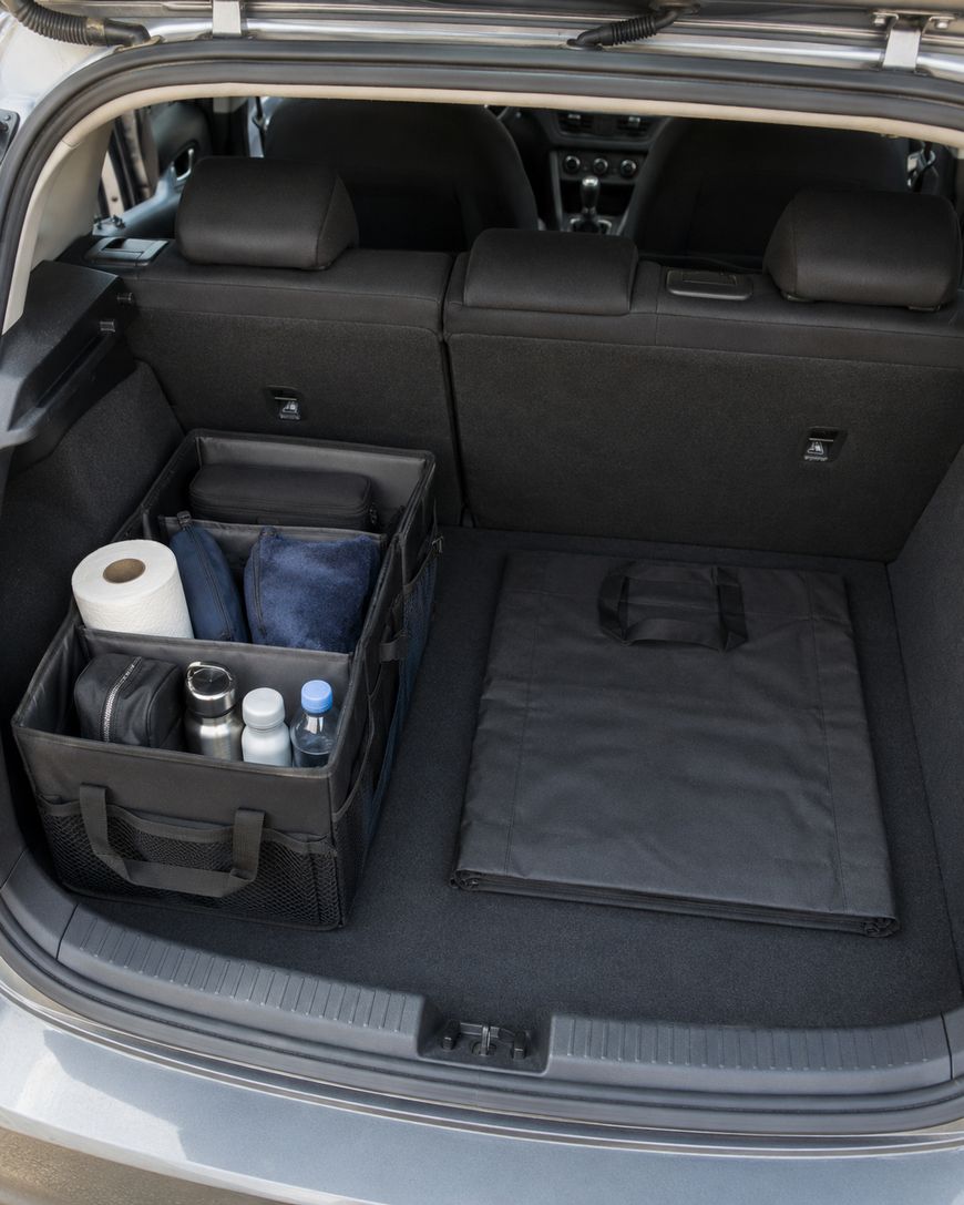 An organized car trunk with a black storage bin holding paper towels, bottles, and other items next to a folded black mat, with the back seats visible.