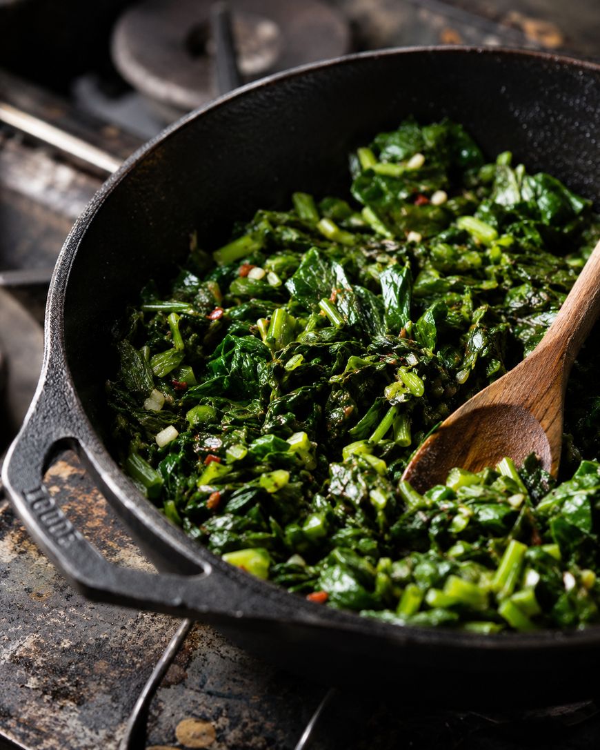 Chopped leafy greens being sautéed in a black cast iron skillet with a wooden spoon, on a stovetop.