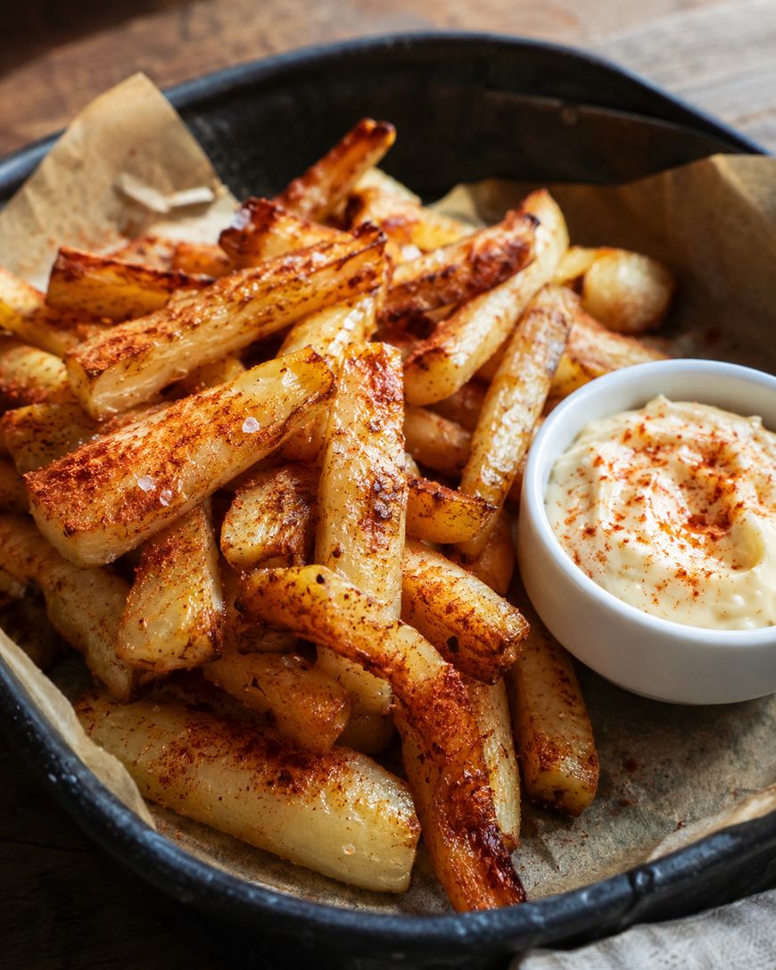A basket filled with seasoned, baked fries sprinkled with paprika, served on parchment paper next to a small dish of creamy dipping sauce.