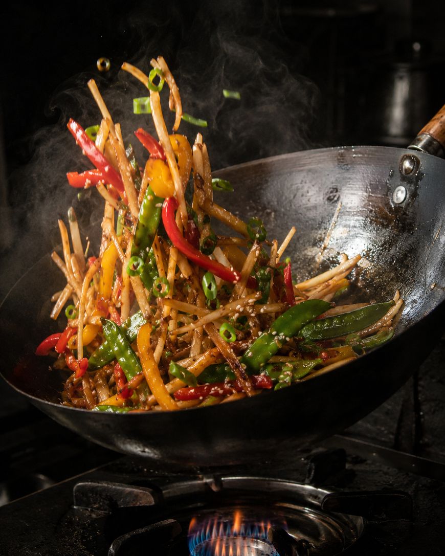Colorful stir-fried vegetables, including bell peppers, green beans, and bean sprouts, are being tossed in a wok over a gas flame, with steam rising from the sizzling ingredients.