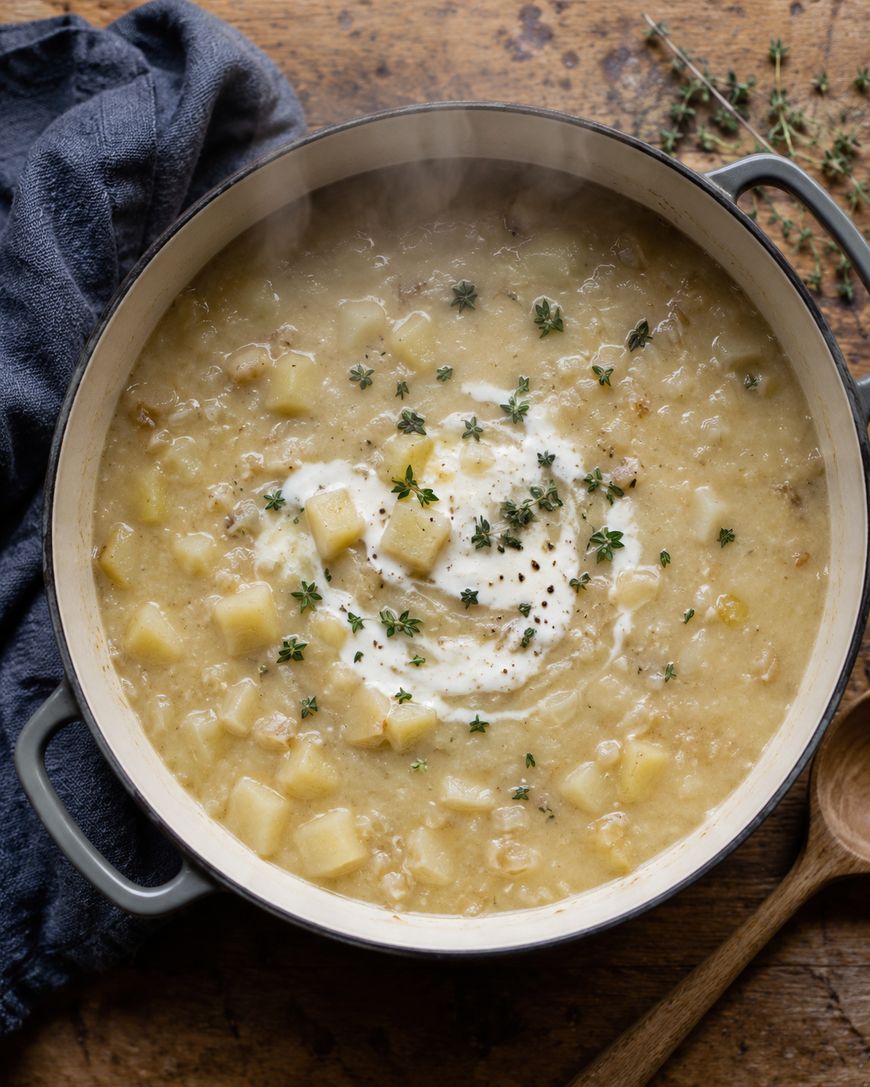 A pot of creamy potato soup garnished with a swirl of cream, cracked black pepper, and fresh thyme, sits on a wooden surface next to a wooden spoon and a dark cloth napkin. Steam rises from the hot soup.