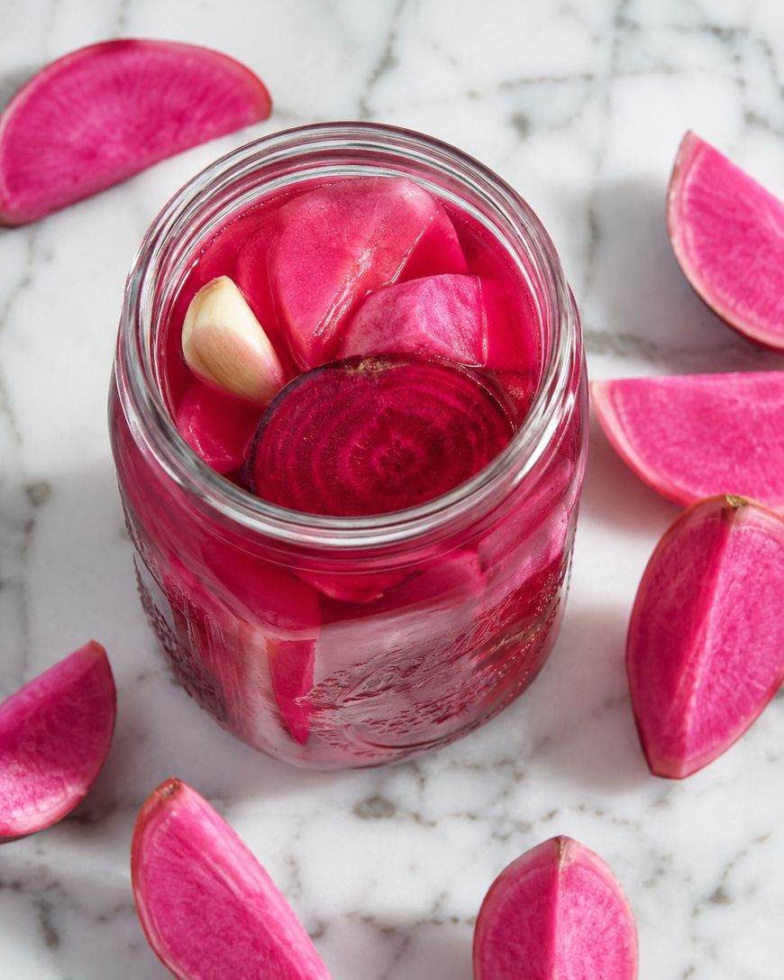 A glass jar filled with vibrant pink pickled radishes and a clove of garlic, surrounded by fresh radish wedges on a white marble surface.
