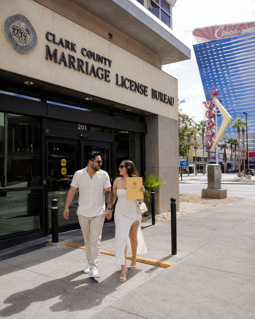 A couple dressed in light-colored clothes smiles while walking out of the Clark County Marriage License Bureau in Las Vegas, holding an official envelope. The Vegas Strip and the Fremont Street sign are visible in the background.