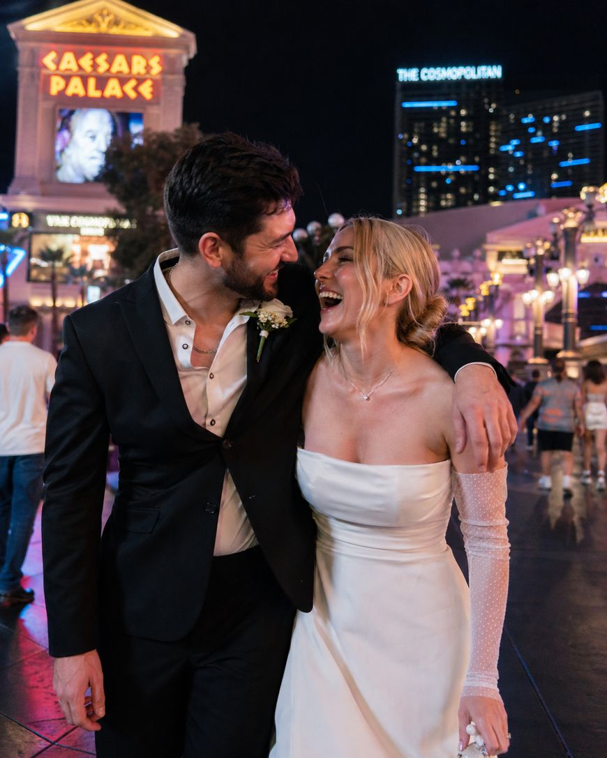 A smiling couple, dressed in wedding attire, walk arm-in-arm at night on a lively street. Bright signs for Caesars Palace and The Cosmopolitan are visible in the background.