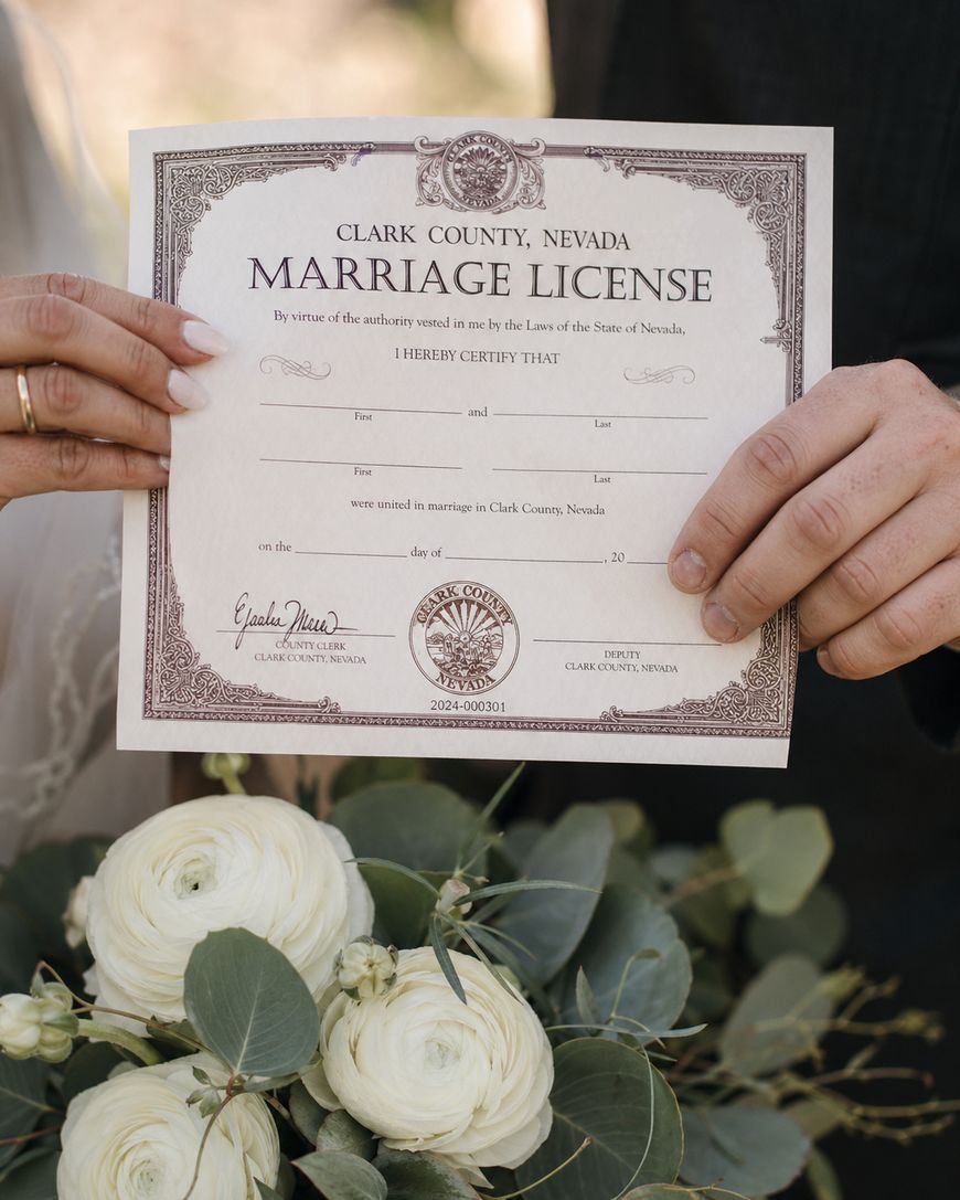 Two people hold a Clark County, Nevada marriage license together above a bouquet of white flowers and greenery. Their faces are not visible, only their hands and part of their outfits can be seen.