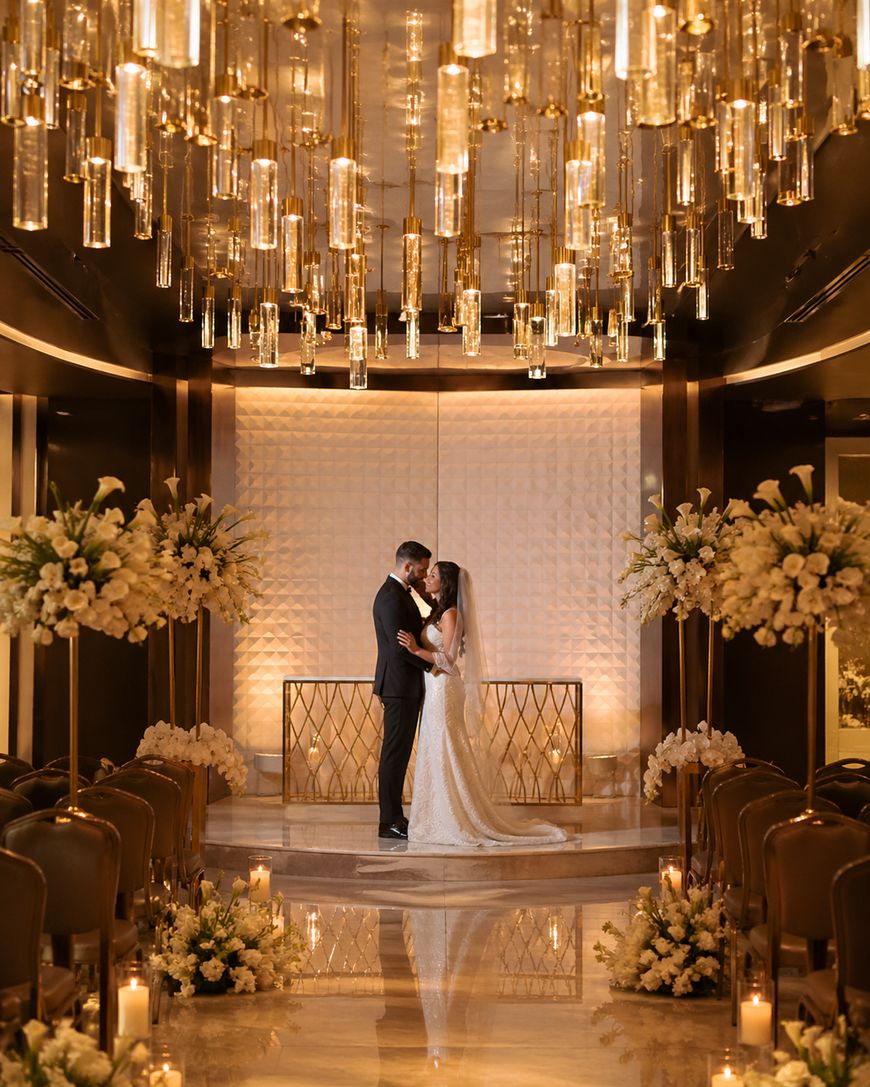 A bride and groom stand embracing at the end of an aisle decorated with white flowers under a dramatic golden chandelier in an elegant, modern indoor wedding venue.