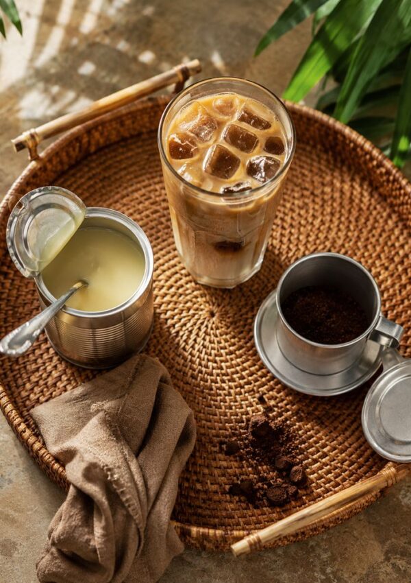 A woven tray holds a glass of Ca Phe Sua Da, an open can of condensed milk with a spoon, a metal coffee filter with ground coffee, a brown cloth napkin, and a few leaves in the background—perfect for your Vietnamese Coffee Recipe.