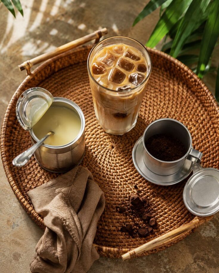 A woven tray holds a glass of Ca Phe Sua Da, an open can of condensed milk with a spoon, a metal coffee filter with ground coffee, a brown cloth napkin, and a few leaves in the background—perfect for your Vietnamese Coffee Recipe.