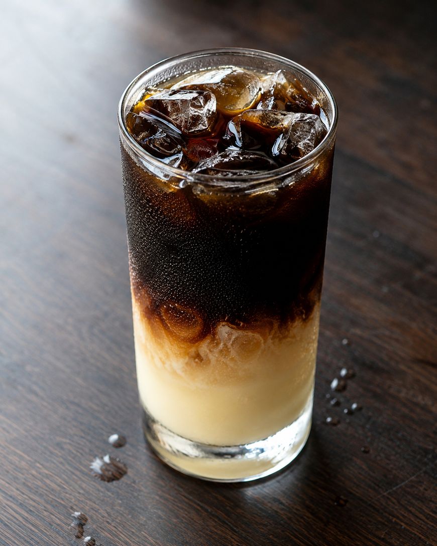 A tall glass filled with iced coffee, featuring distinct layers of dark coffee on top and creamy milk at the bottom, sits on a dark wooden surface with some condensation and water droplets around it.