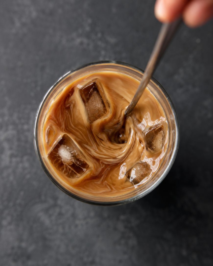 A hand stirs a glass of iced coffee with a metal spoon, creating swirling patterns as the milk mixes with the coffee. Several ice cubes float in the drink, and the background is a dark textured surface.