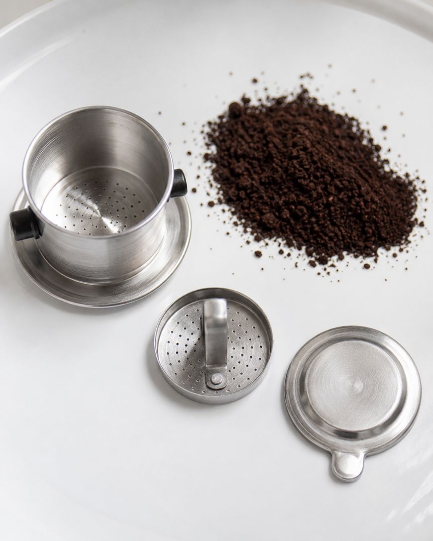 A stainless steel Vietnamese coffee filter disassembled into three parts sits on a white surface next to a pile of ground coffee.