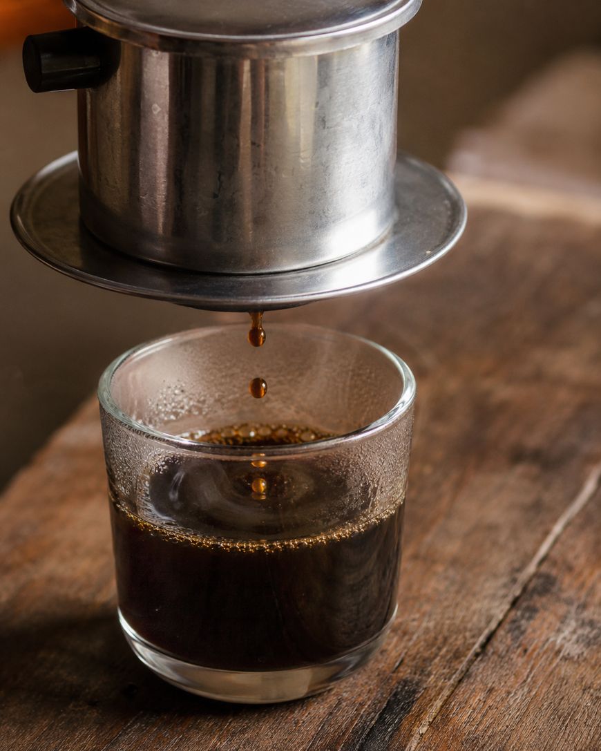 A metal Vietnamese coffee filter drips coffee into a glass cup on a rustic wooden surface.