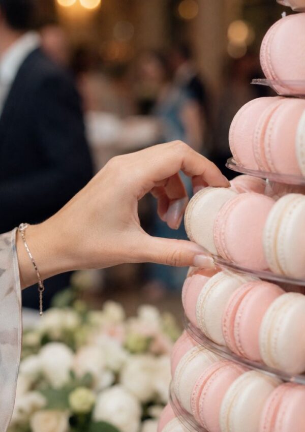 A hand reaches to pick a pink macaron from a tiered display of pink and white macarons—an elegant wedding dessert idea that goes beyond traditional cake, with a blurred background of people and flowers.