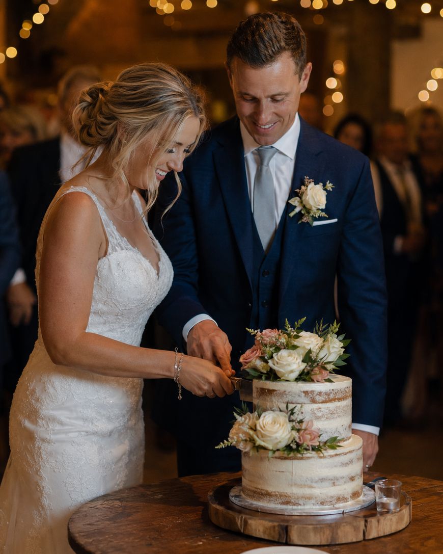 A bride and groom smile as they cut a two-tiered, semi-naked wedding cake decorated with white and blush flowers, standing together in a warmly lit venue with guests blurred in the background.