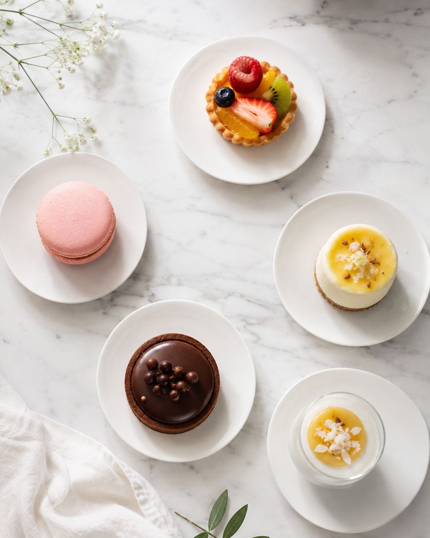 Five assorted desserts on white plates, including a pink macaron, fruit tart, chocolate tart, mini cheesecake, and a creamy dessert in a glass, arranged on a marble surface with floral decoration.
