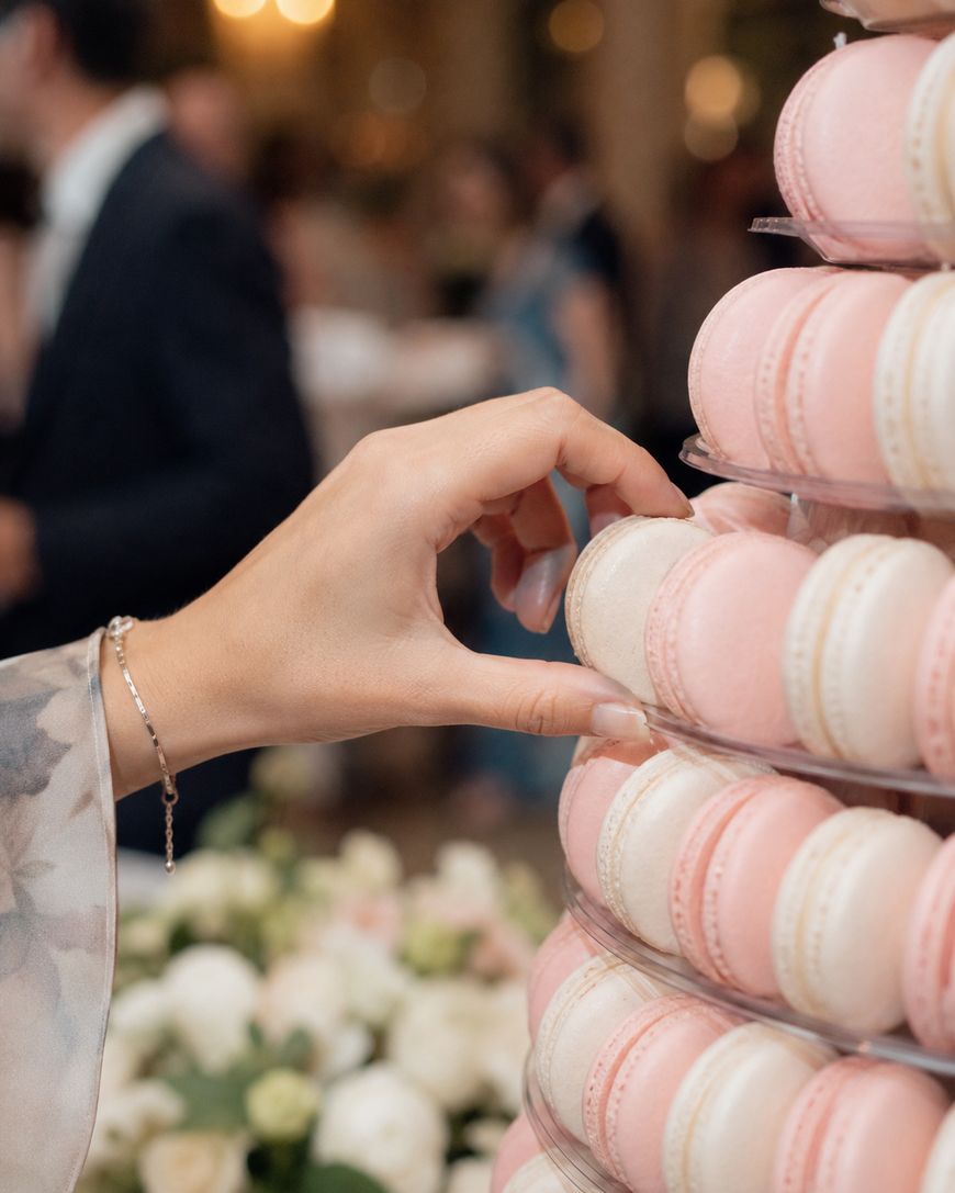A hand reaches for a pink macaron from a tiered display of pink and white macarons at an elegant event, with blurred guests and floral arrangements in the background.