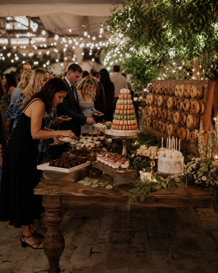 Guests in formal attire select desserts from a rustic wooden table filled with pastries, cakes, and a macaron tower, with a donut wall and greenery in the background under string lights.