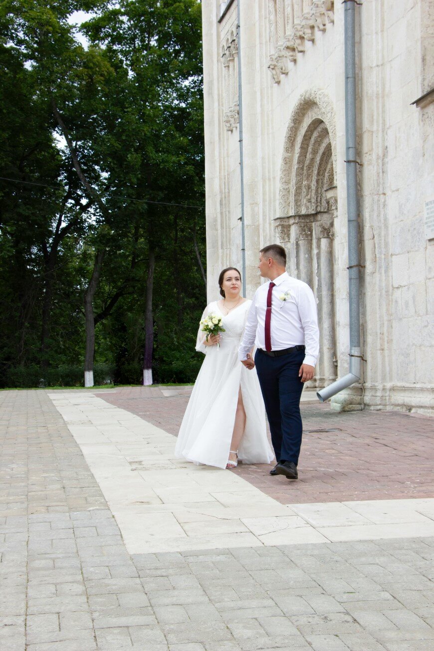 A bride and groom walk hand-in-hand on a paved path in front of a large stone building with arched doorways and carved details.