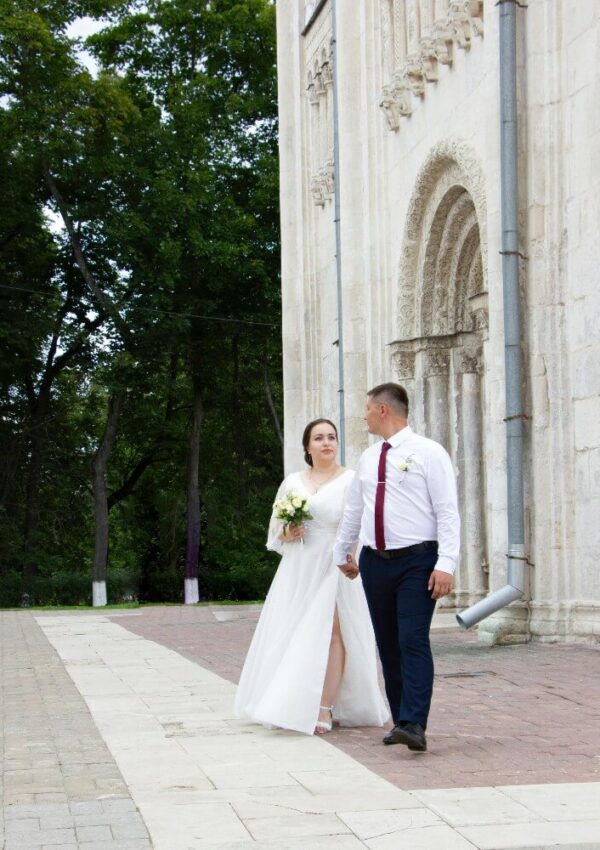 A bride in a white dress and groom in a white shirt and dark pants walk hand-in-hand past a stone building with an arched entrance.