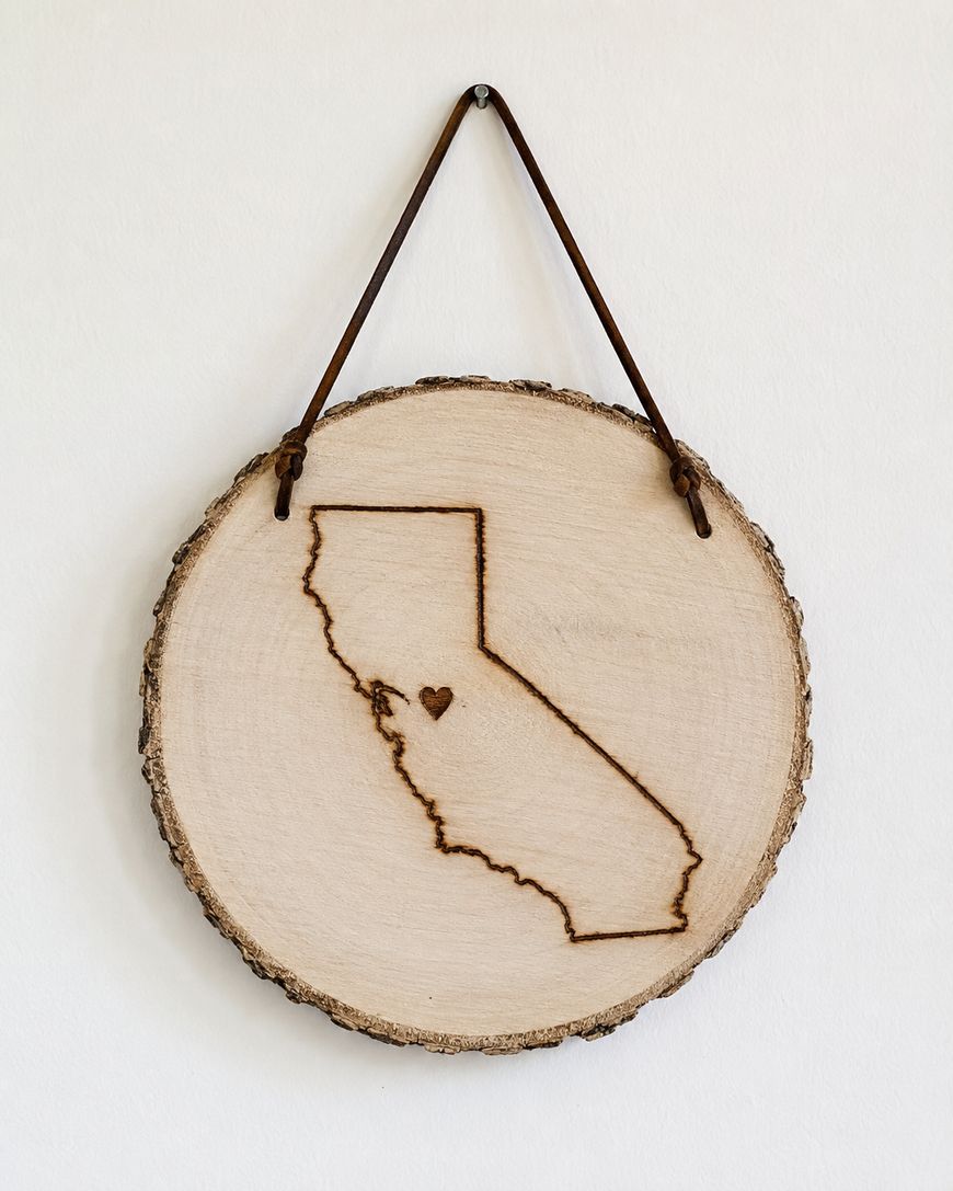 A round wooden plaque with a leather strap displays a burned outline of California and a small heart near the center of the state.