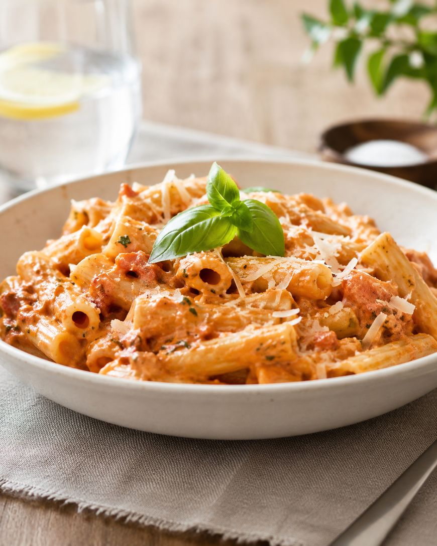 A bowl of creamy pasta topped with grated cheese and a fresh basil leaf, served on a neutral napkin with a glass of water and a plant in the blurred background.