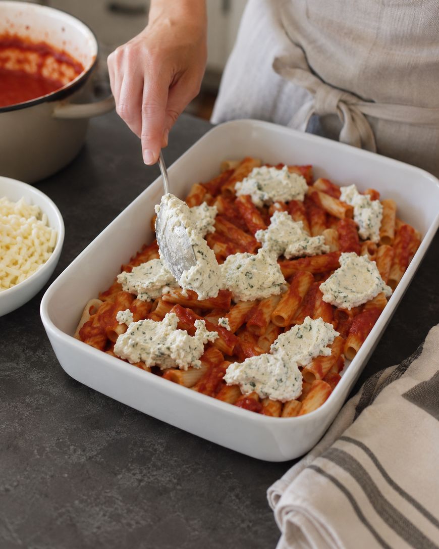 A person spreads ricotta cheese mixture over a layer of baked ziti pasta with tomato sauce in a white casserole dish. Nearby are a pot of sauce and a bowl of shredded cheese on a dark countertop.