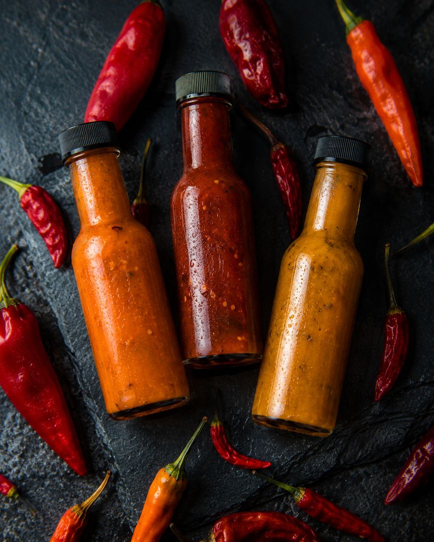 Three bottles of hot sauce in varying shades of orange and red are arranged on a dark surface, surrounded by whole red chili peppers.