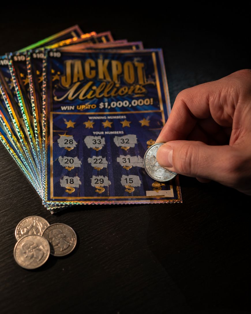 A hand uses a coin to scratch off a Jackpot Millions lottery ticket on a dark surface, with several other scratch tickets and coins nearby.