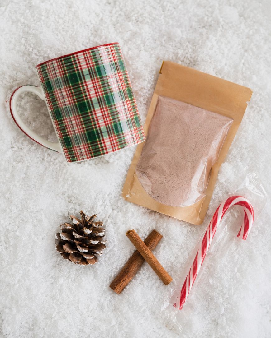 A plaid mug, a pouch of hot cocoa mix, a wrapped candy cane, a pinecone, and two cinnamon sticks arranged on a snowy surface.